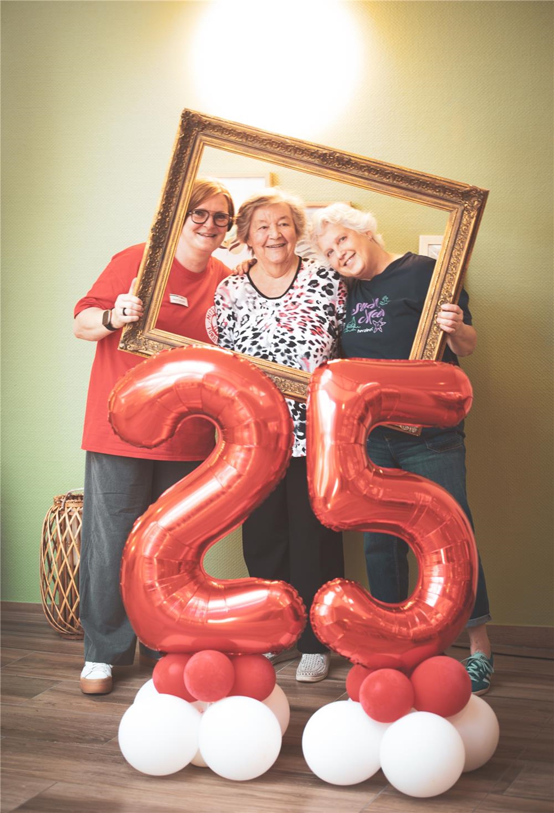 Drei Mitarbeitende schauen fröhlich durch einen goldenen Rahmen, vorne zwei große rote Ballons mit einer 25 